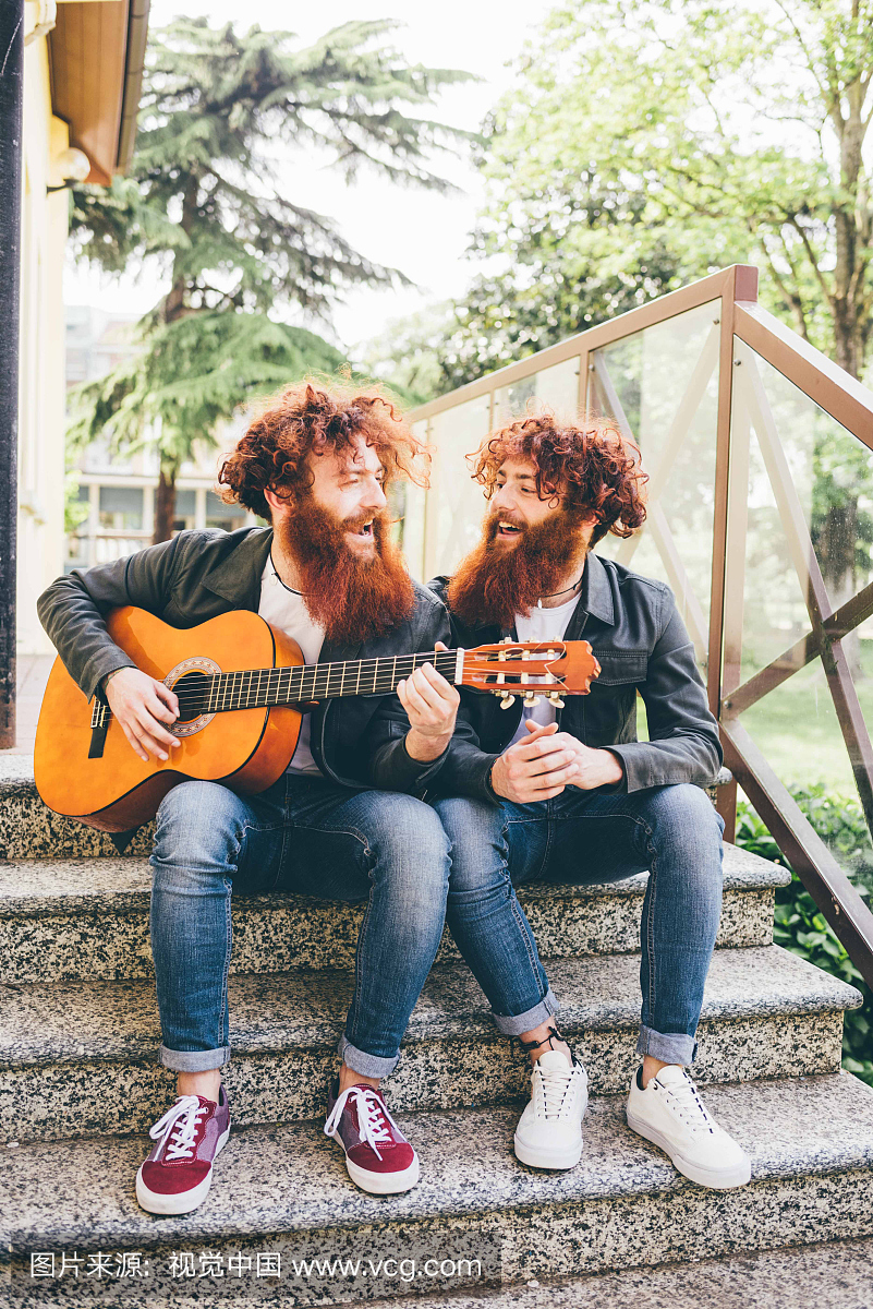年轻男性hipster双胞胎与红胡子坐在楼梯弹吉他