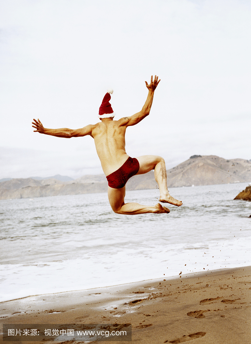穿圣诞老人帽子和红色泳裤短裤跳在海滩,后视
