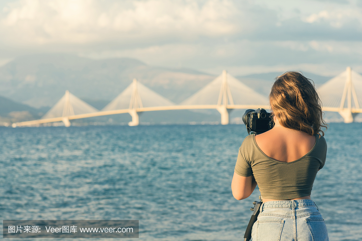 年轻女性在短裤和一件T恤在海滩上与三脚架和相机拍摄的希腊帕特雷附近的Rion-Antirion桥的照片