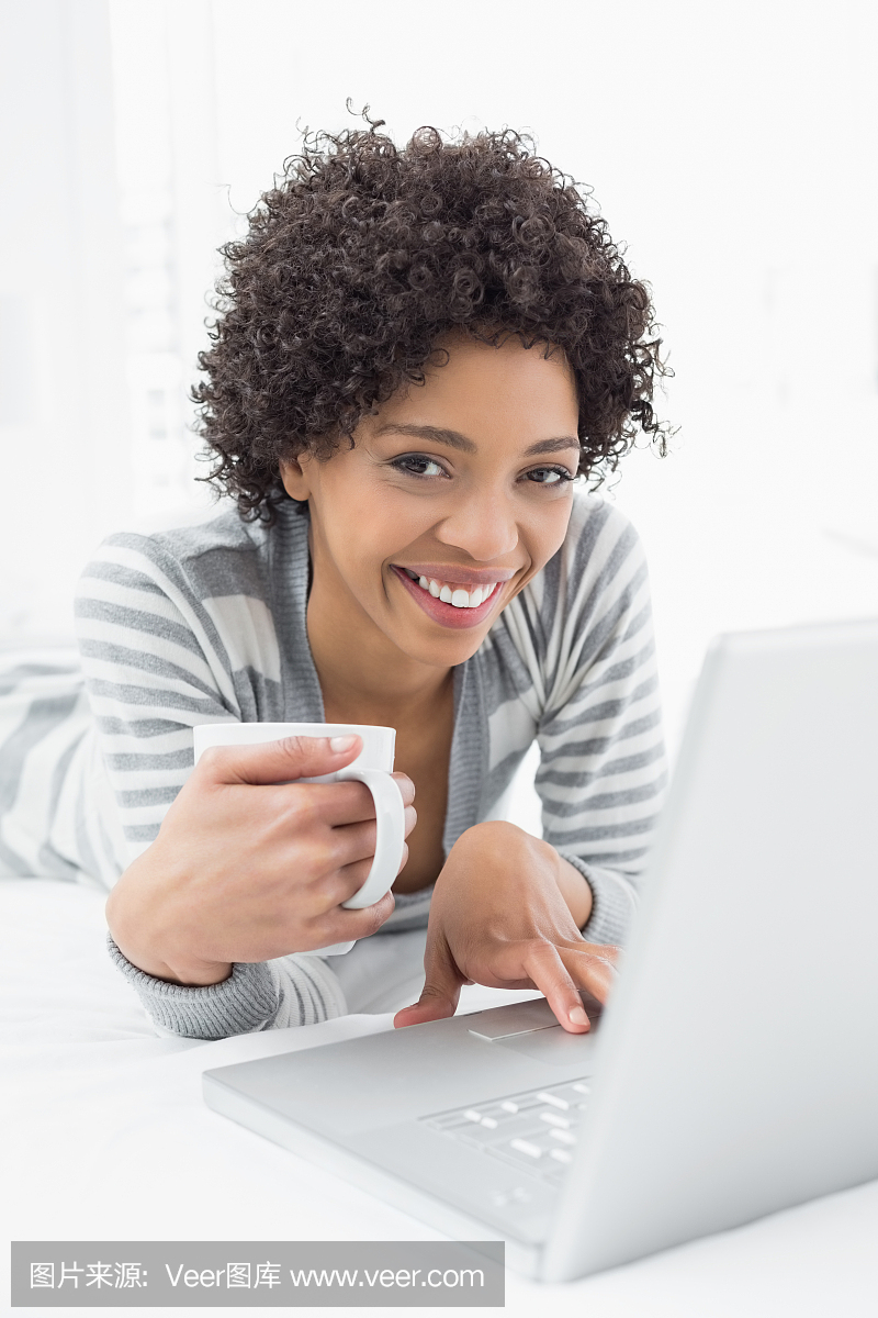 微笑与咖啡杯在床上使用笔记本电脑的女人
