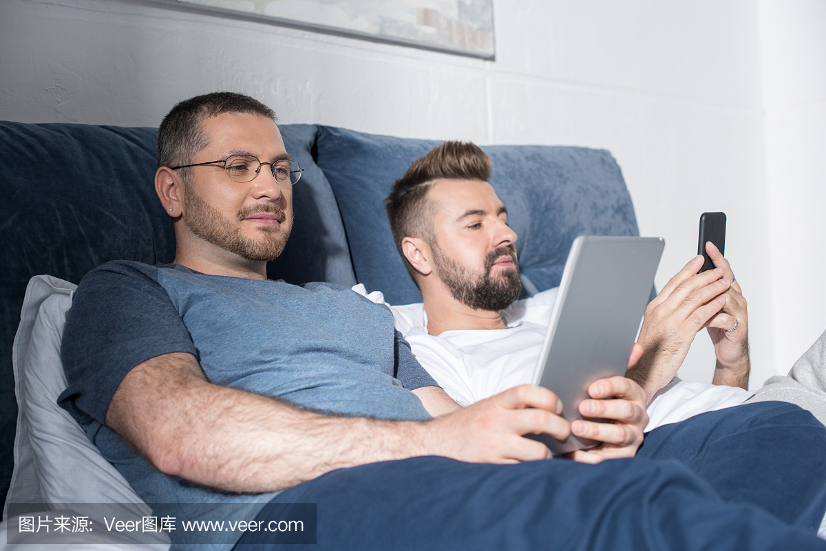 两个有胡子的男人躺在床上和使用数字设备