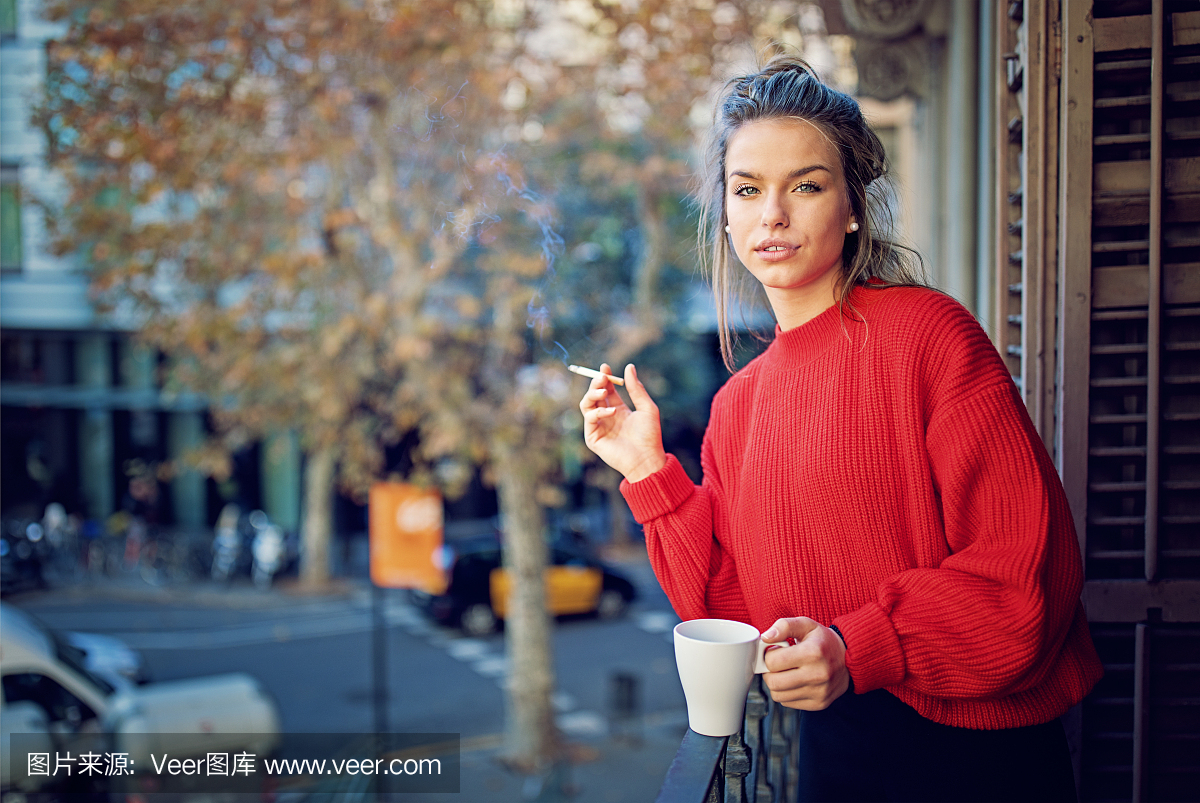 年轻的女孩在早晨吸烟,在阳台喝咖啡