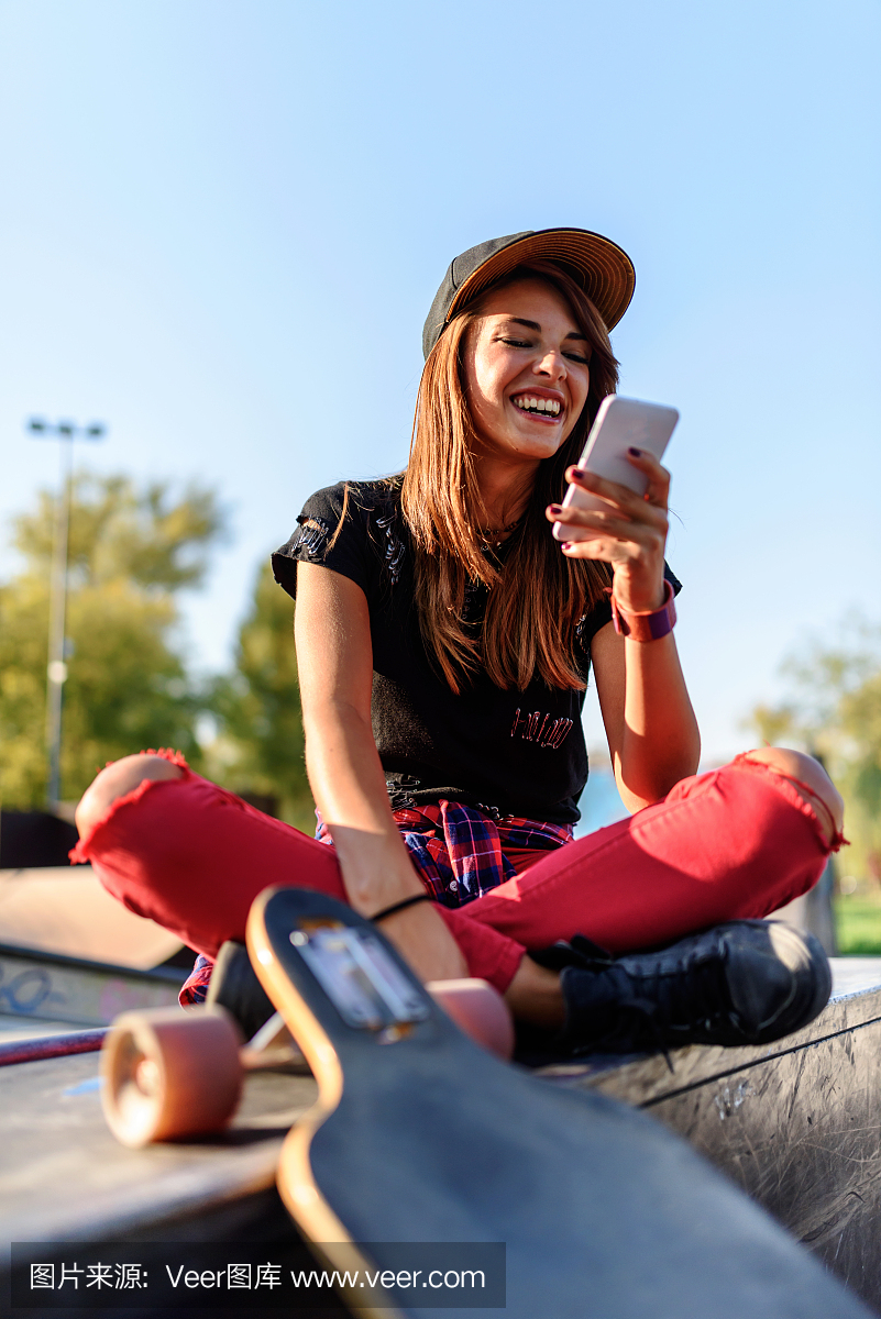 可爱的城市女孩在滑板公园与使用智能手机的滑