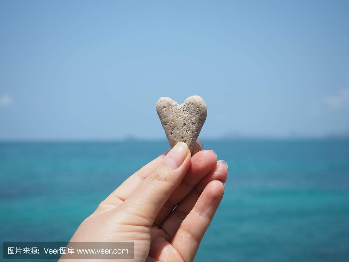 拿着心脏形状的石头在蓝色夏天沙滩背景的女人