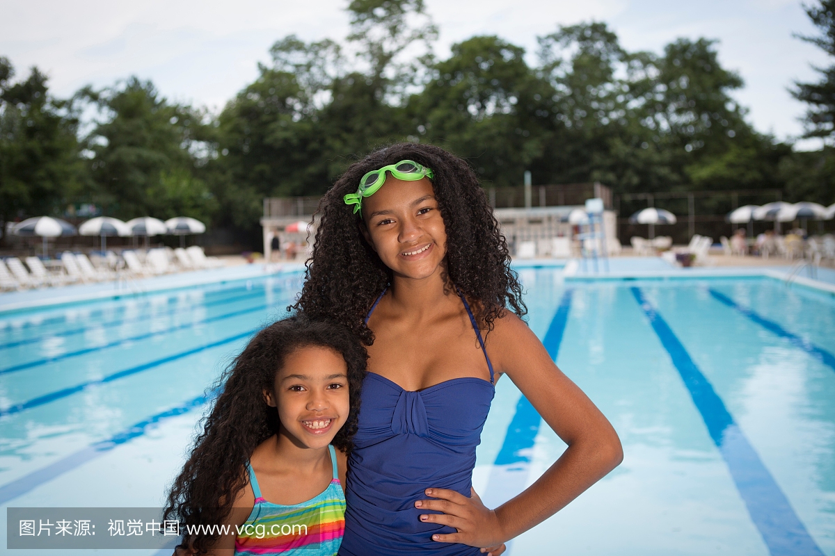 穿泳装的姐妹站在游泳池前面,看着相机微笑