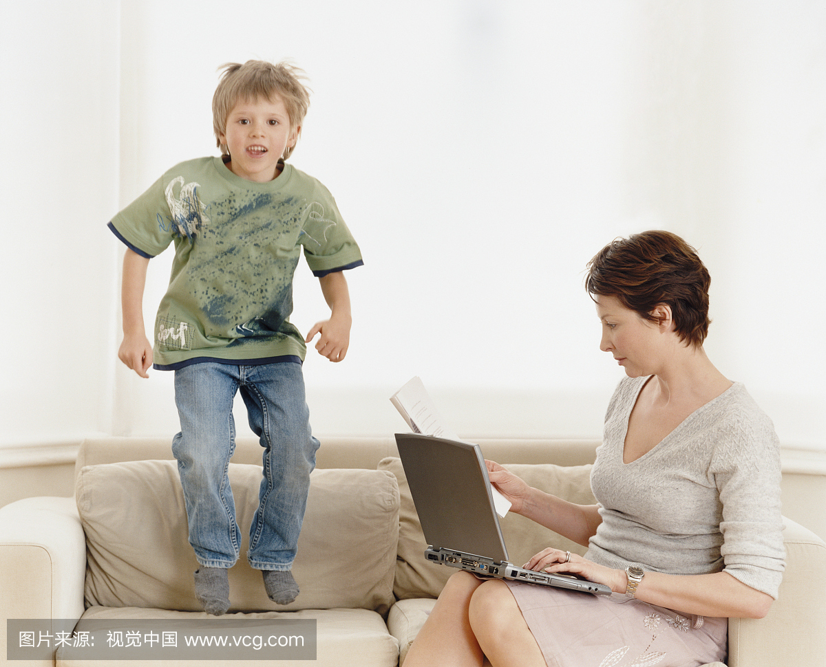 年轻的男孩跳上沙发,而他的母亲在她的笔记本