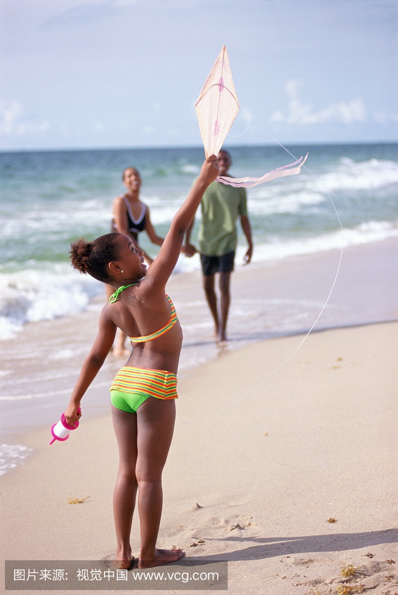 女孩(5-7)放风筝在海滩,后视图,父母在背景中