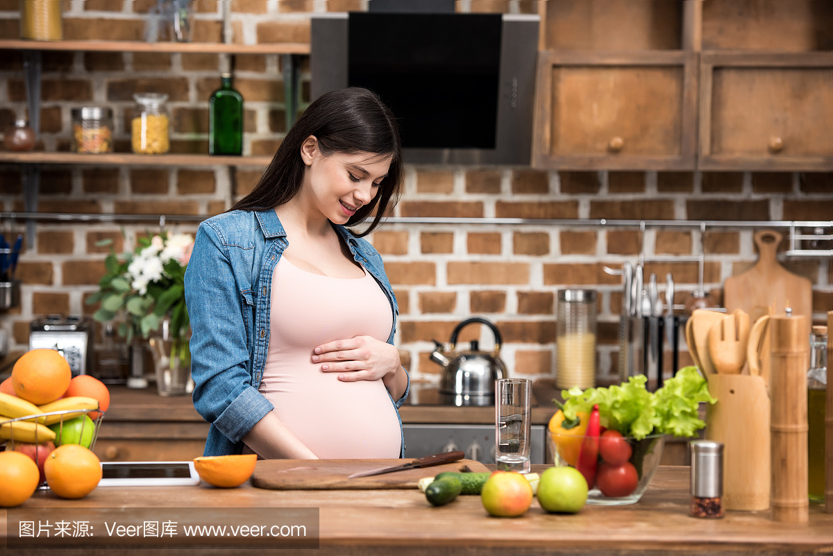 美丽的微笑年轻孕妇摸她的肚子,同时在厨房里